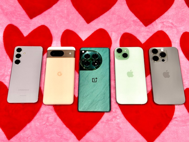 Gray Samsung Galaxy S24 (left), Rose Gold Google Pixel 8, Flowy Emerald OnePlus 12, Green iPhone 15, Titanium Gray iPhone 15 Pro on a pink and red heart blanket.