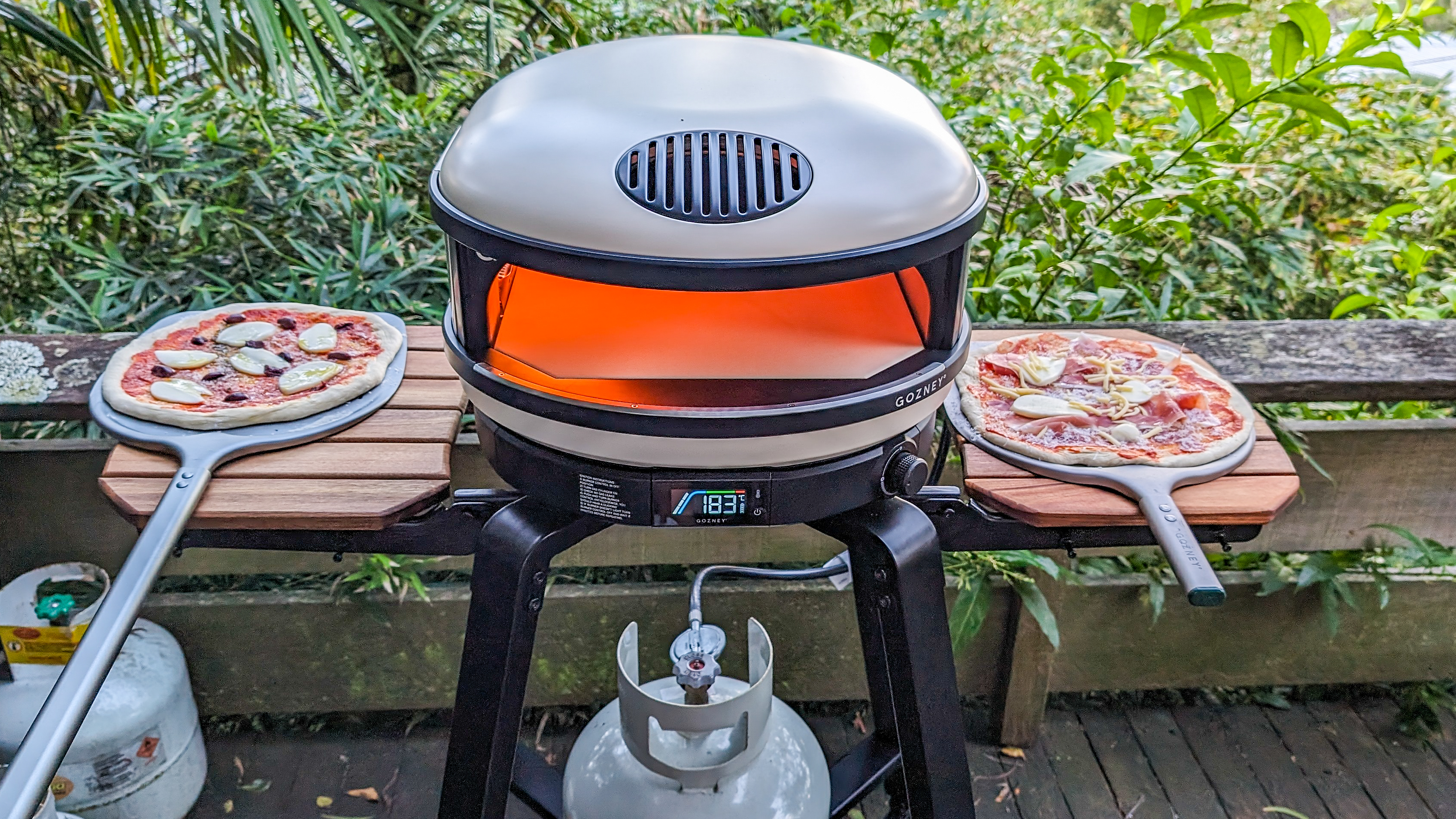 Gozney Arc XL fired up with two uncooked pizzas beside it on the fold-up shelves of its stand