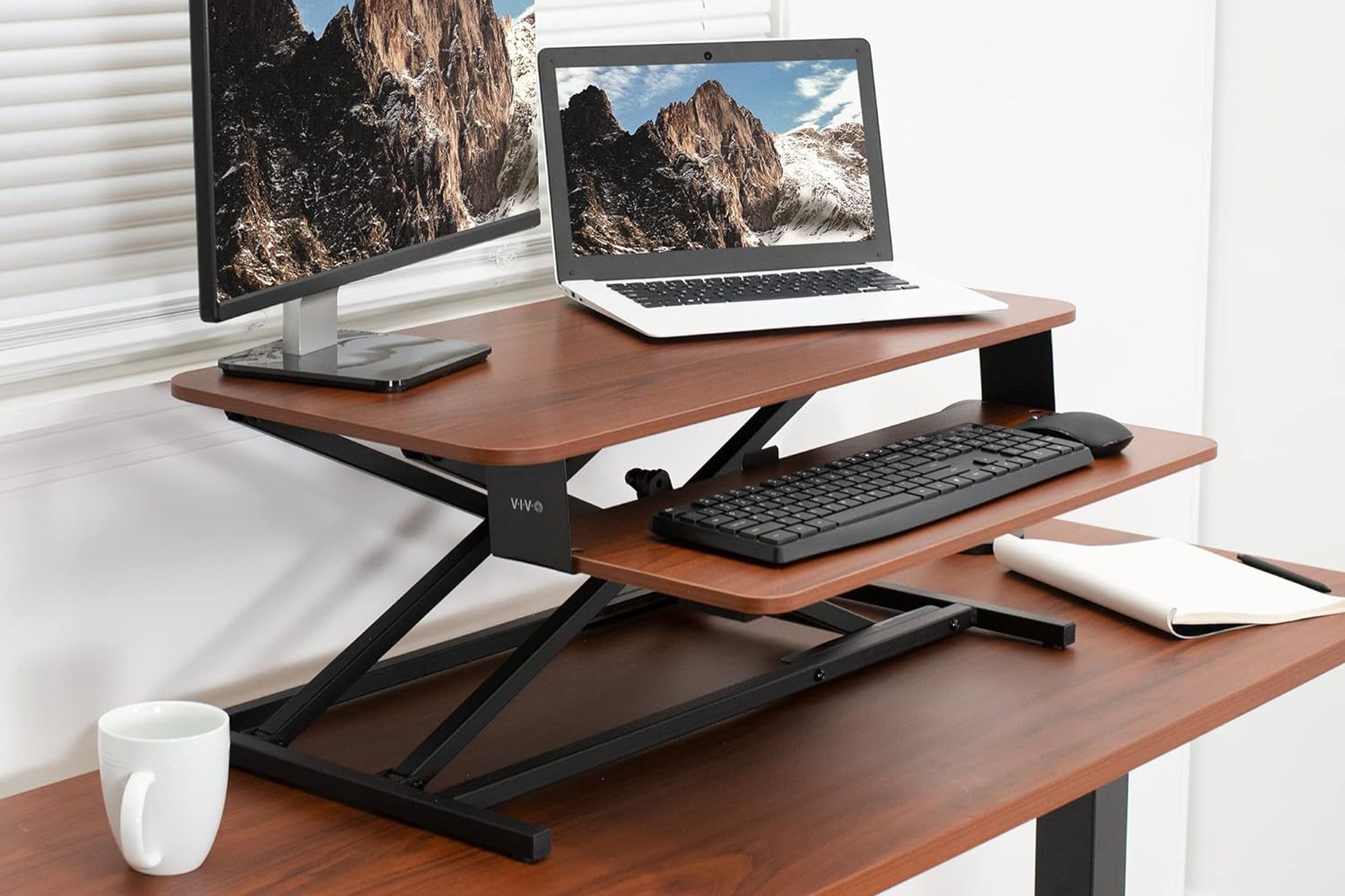 A two-level stand on top of a desk holding a monitor, a laptop, and a keyboard.