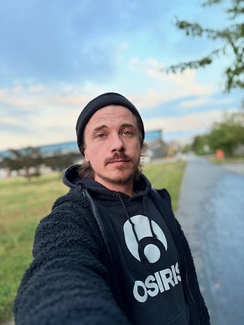 Eine Person mit Bart, die einen schwarzen Hoodie und eine Mütze trägt und draußen mit einem bewölkten Himmel ein Selfie macht.