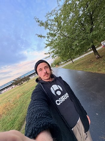 Ein Mann macht ein Selfie in einem Park mit Bäumen und einem bewölkten Himmel im Hintergrund.