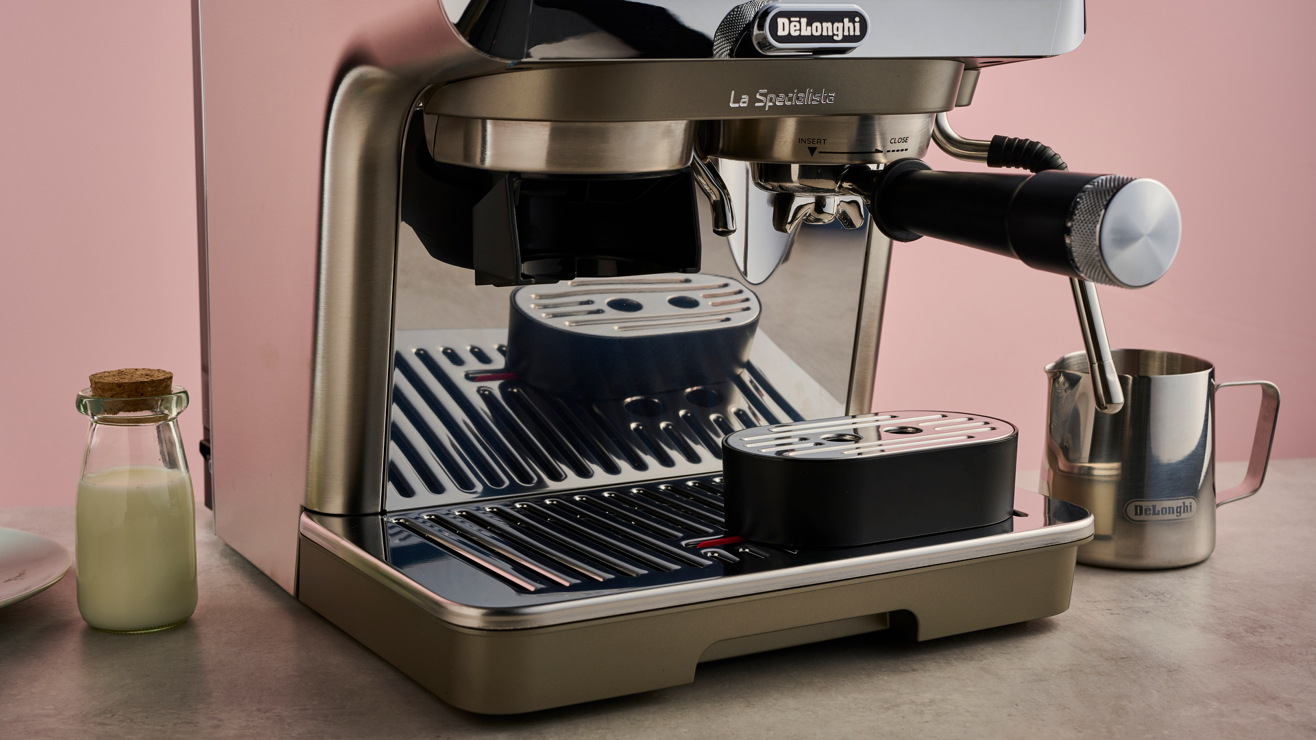 The De'Longhi Specialista Arte Evo sits on a stone-effect surface in front of a pink backdrop. It has a small cork-topped bottle of milk to its left, and a metal milk jug to its right.