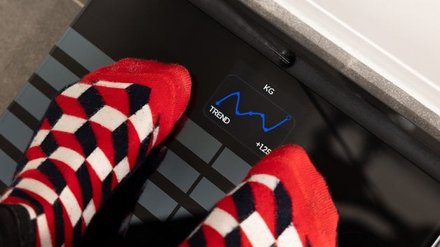 Feet in patterned socks on a Withings Body Scan scale displaying weight and trend graph.