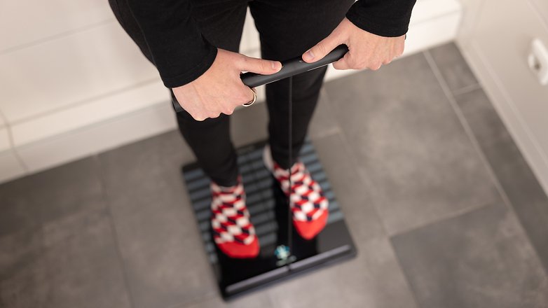 Person standing on a Withings Body Scan Smart Scale, holding the handle, wearing red and black checkered socks.