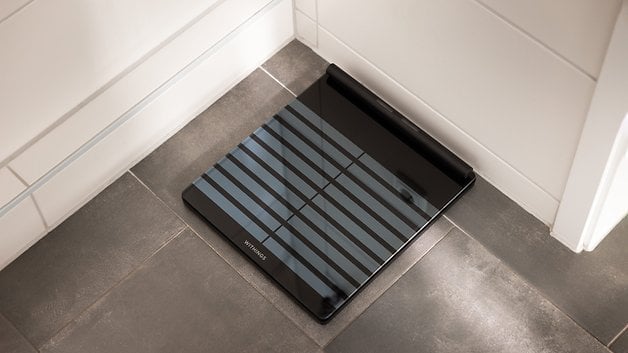 A Withings Body Scan smart scale on a tiled floor in a bathroom.